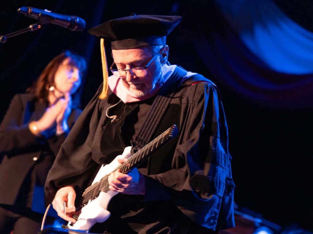 Person in graduation cap and gown playing electric guitar on stage, with another person clapping in the background.