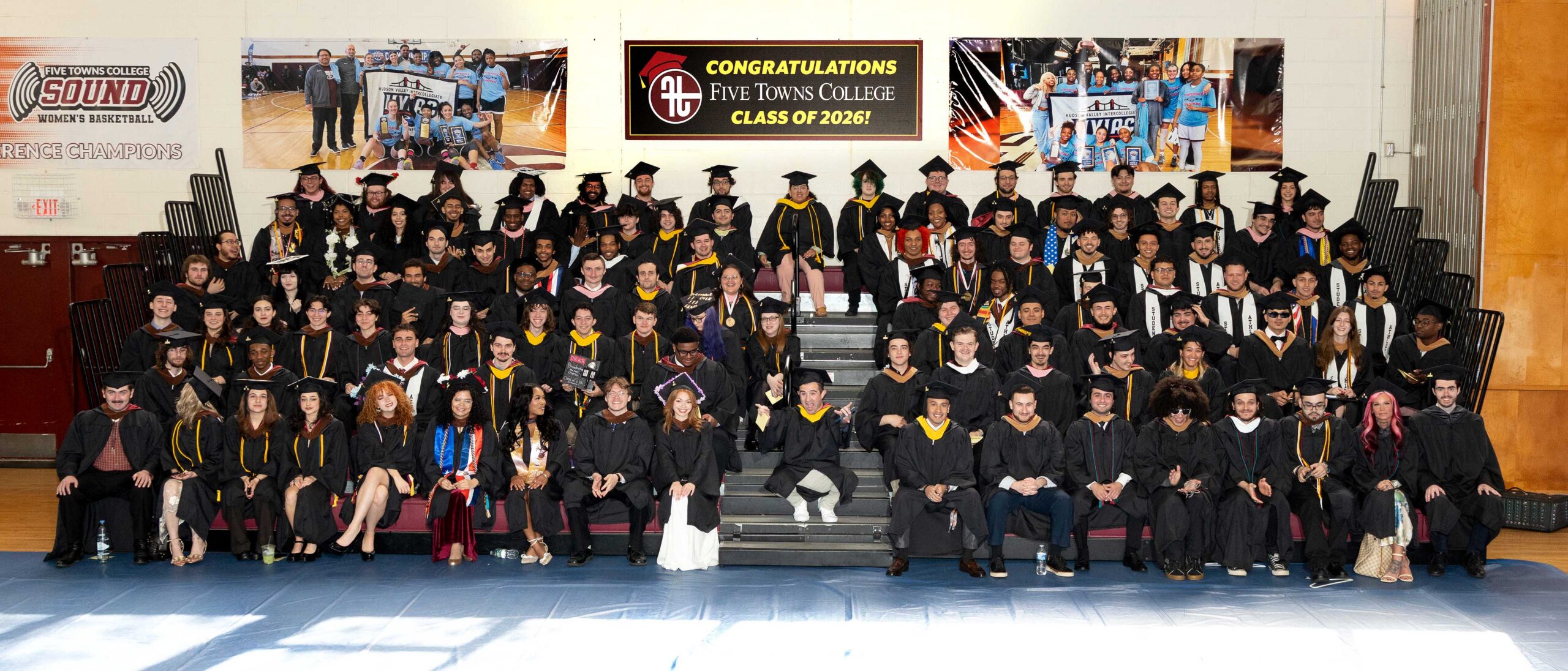 A large group of creative college graduates in caps and gowns pose for a class photo on bleachers in a gymnasium. A banner above reads Congratulations Five Towns College Class of 2026!.