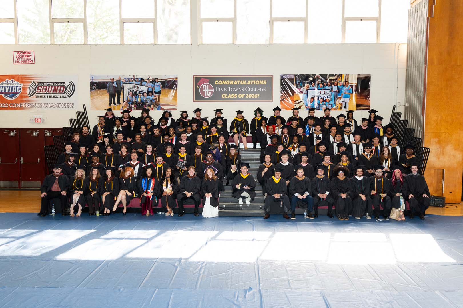 A large group of graduates in caps and gowns pose for a group photo indoors, seated and standing on bleachers in front of a congratulatory banner for the Five Towns College Class of 2022.