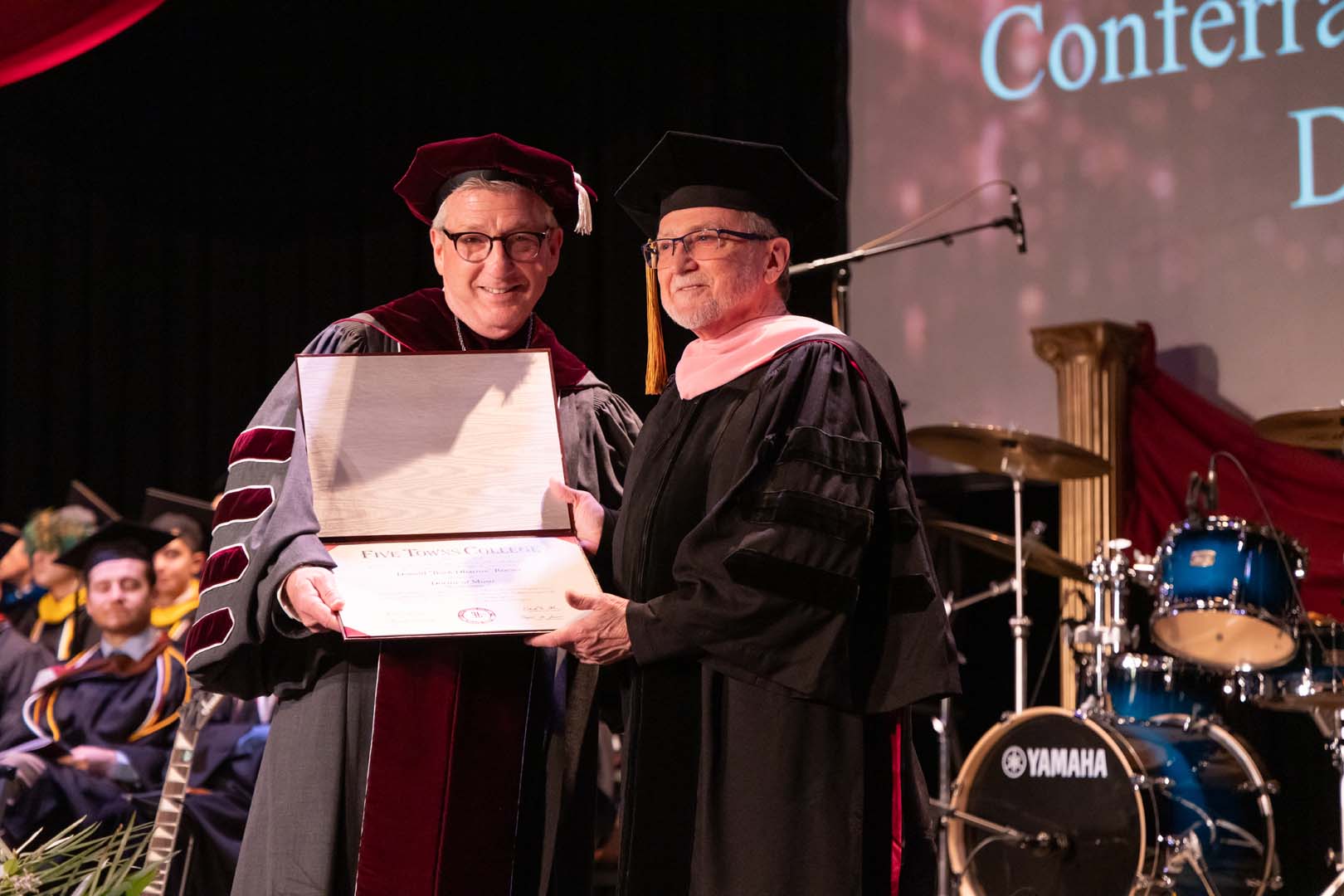 Two men in academic regalia smile and hold a diploma together on stage during a graduation ceremony. In the background, musicians in caps and gowns sit near a drum set, with a graduation banner projected on the wall.