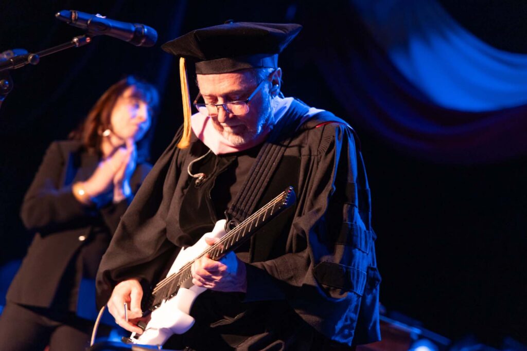 A person in academic regalia plays an electric guitar on stage under colorful lighting, while another person in the background claps along.