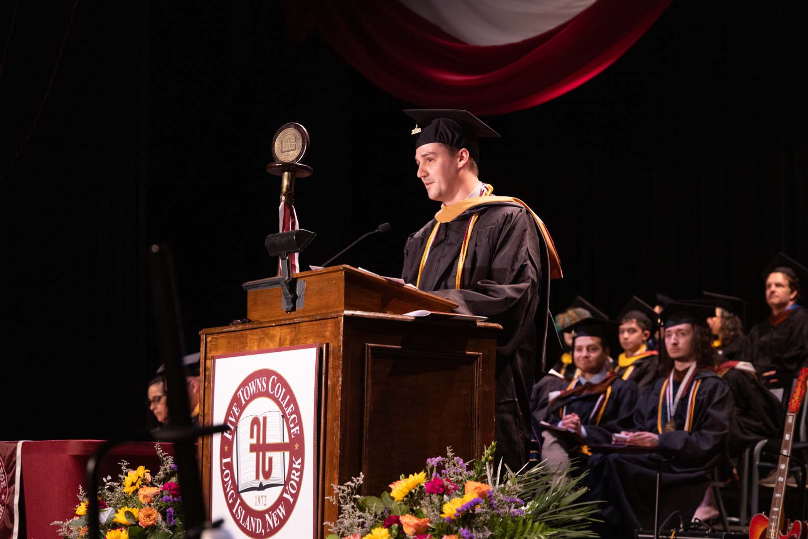 A graduate in cap and gown stands at a podium delivering a speech during a graduation ceremony. Other graduates sit on stage behind him. The stage is decorated with flowers and a college emblem.