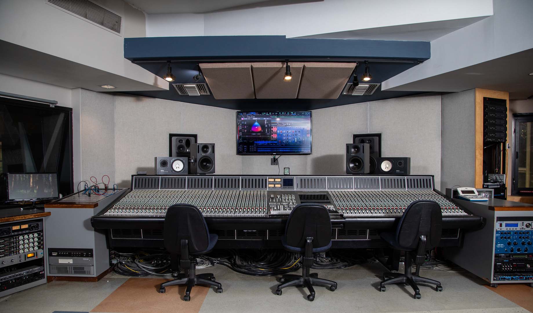 A professional audio recording studio control room with three chairs facing a large mixing console, studio monitors, racks of audio equipment, and a computer screen displaying audio software.
