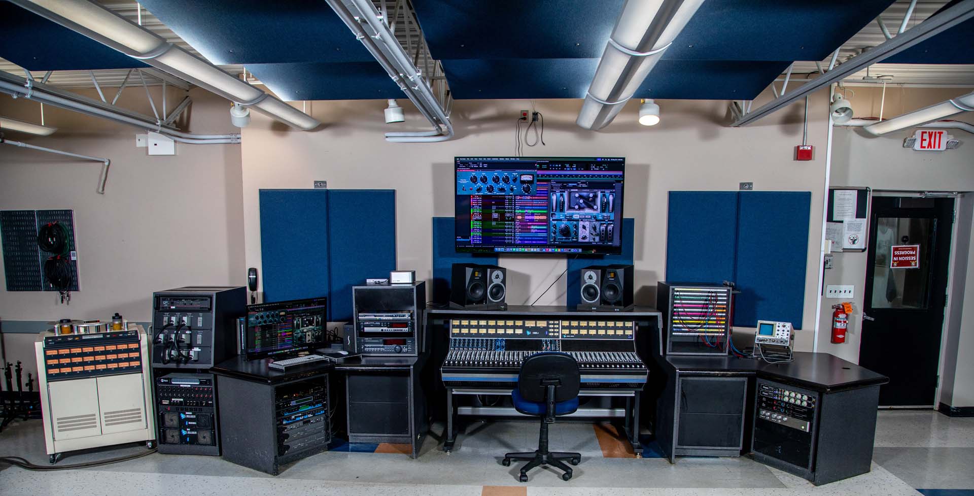 A professional audio recording studio with a large mixing console, multiple audio racks, speakers, a central computer monitor displaying audio software, and various equipment arranged around a black chair.