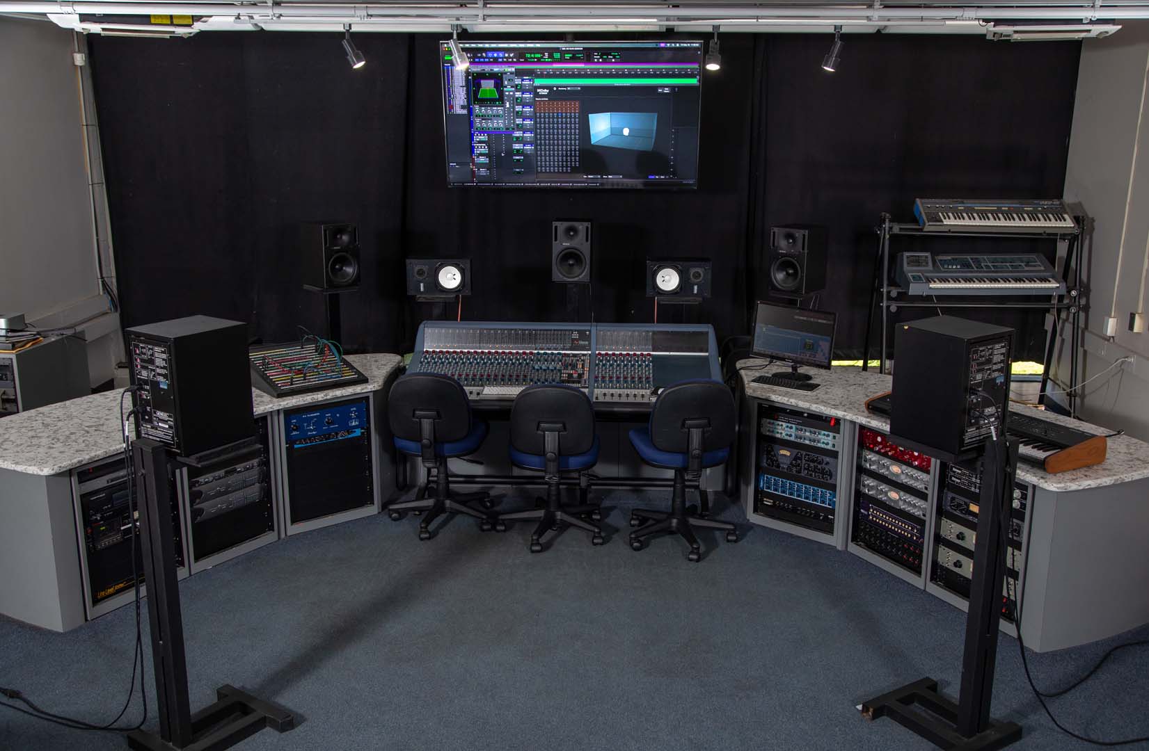 A professional music studio equipped for audio recording, featuring mixing consoles, audio monitors, various sound equipment, three chairs at the main desk, and a large screen displaying audio software on the wall.