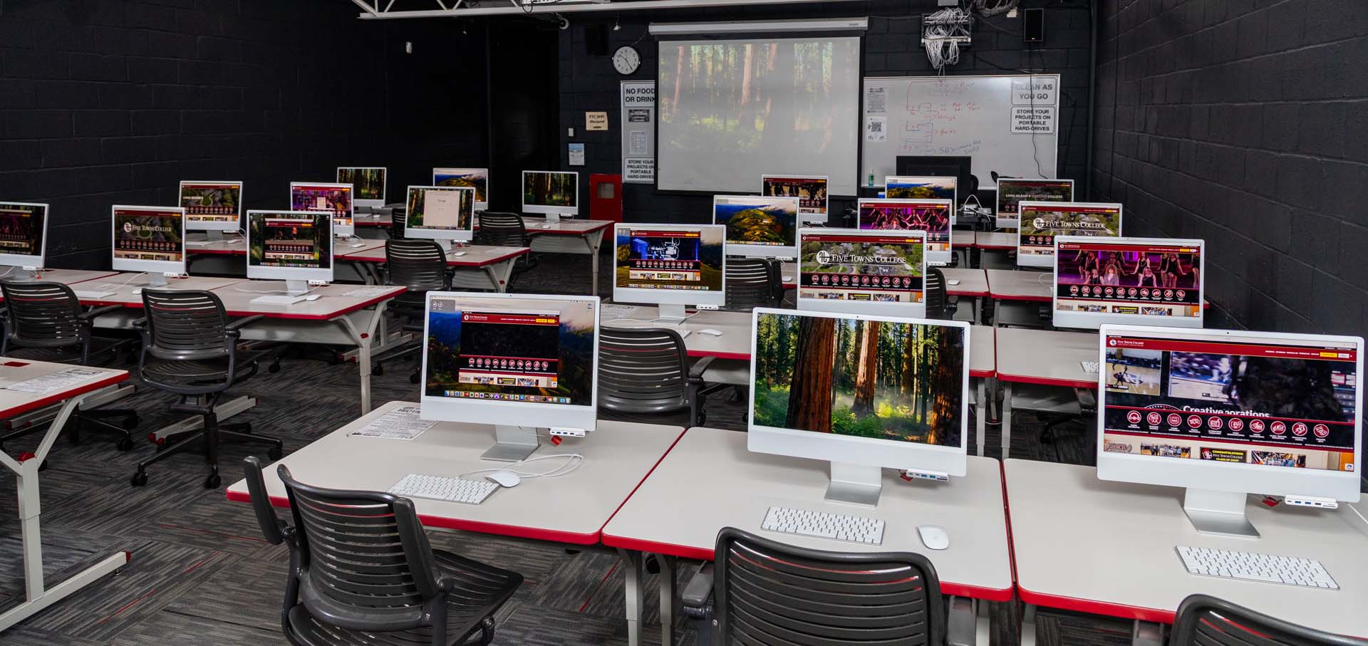 A modern classroom with rows of desks, each equipped with a computer displaying nature scenes. The room has black walls, a large screen at the front, a whiteboard, empty black chairs, and built-in audio recording equipment.