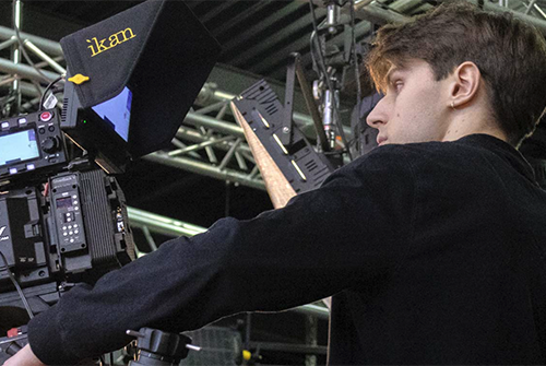 An undergraduate operates a professional video camera with lighting rigs and metal trusses in the background.
