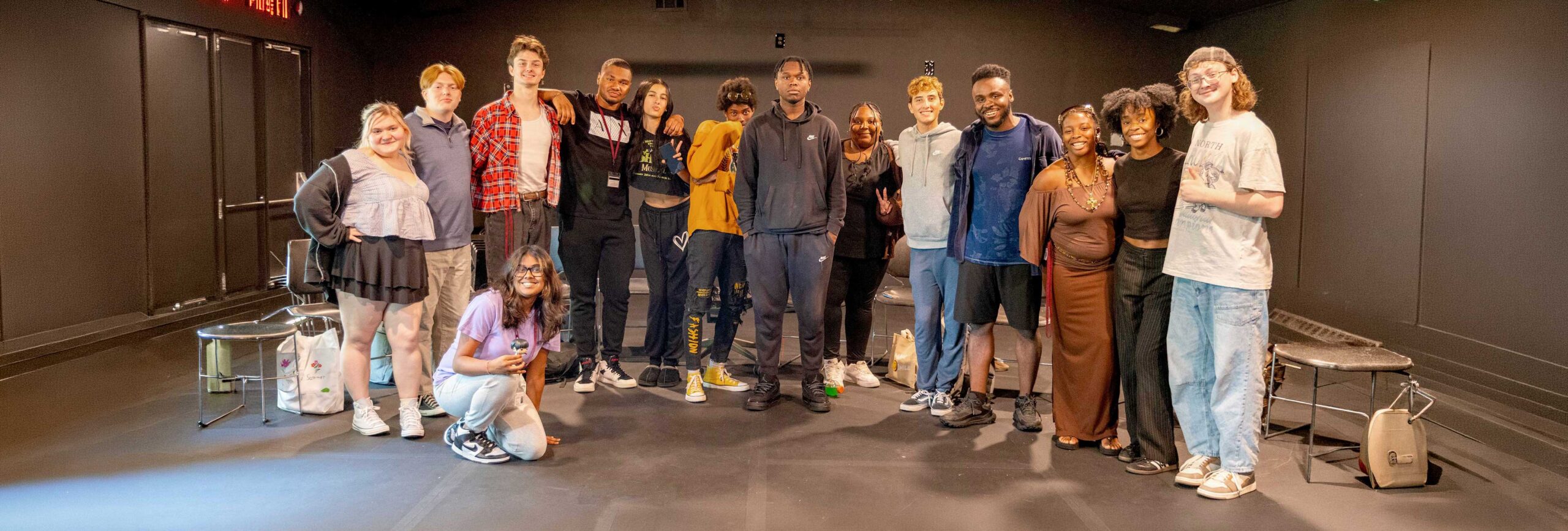 Group of sixteen young adults standing together in a dark room, smiling at the camera.