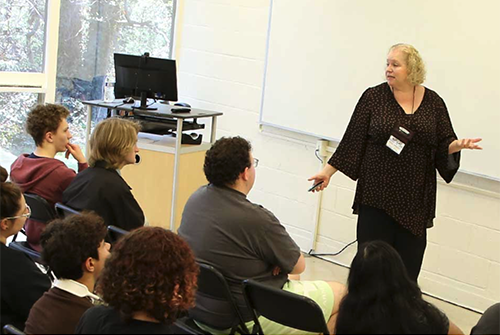 A woman stands and speaks to an undergraduate audience in a classroom, gesturing with one hand.