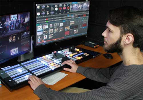 Undergraduate man operating video switcher and editing controls in front of multiple monitors in a production studio.