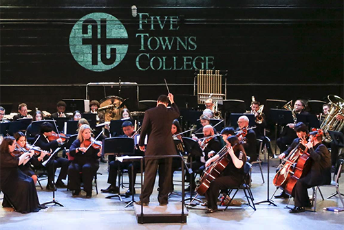 An undergraduate conductor leads an orchestra as the Five Towns College logo shines behind them on stage.