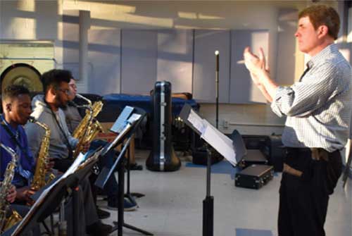 An undergraduate music teacher conducts a group of students playing saxophones in a classroom.