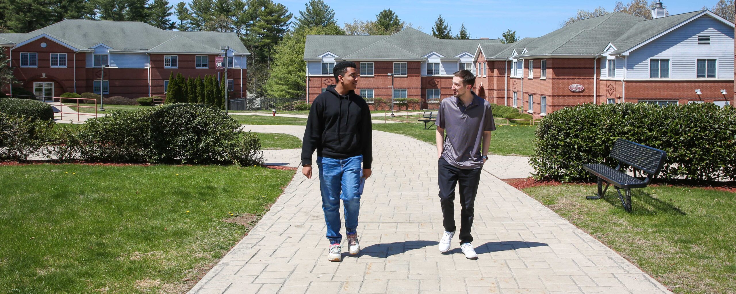Two people walk together on a paved path between red-brick buildings on a sunny day.