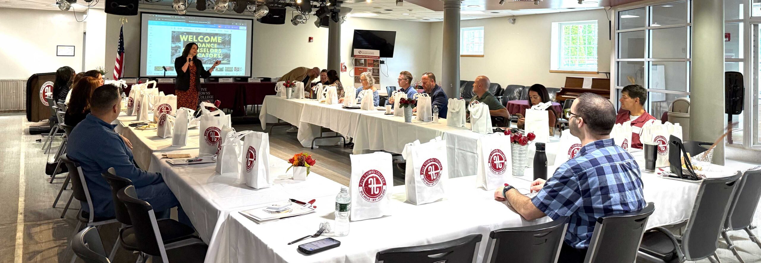 A woman speaks to people seated around tables with gift bags in a bright conference room with a presentation screen.